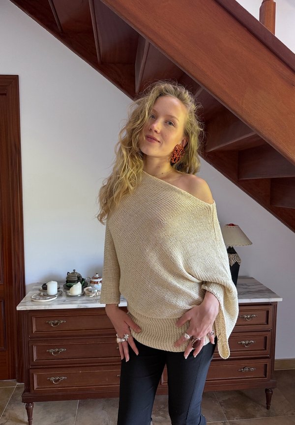 Frau mit langen lockigen blonden Haaren, die einen beigen schulterfreien Pullover, schwarze Hosen, mehrere Ringe und orange Ohrringe trägt, steht drinnen nahe einer Holztreppe.