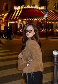 Brun mockajacka och svart klädsel, håller en liten brun handväska. Runda solglasögon. Nattmiljö med kafélampor och randigt markis.