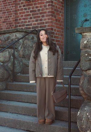 Mujer sonriente con un conjunto informal en tonos beige y marrón, sosteniendo un bolso marrón, de pie sobre escalones de piedra junto a una pared de ladrillos y una puerta verde.