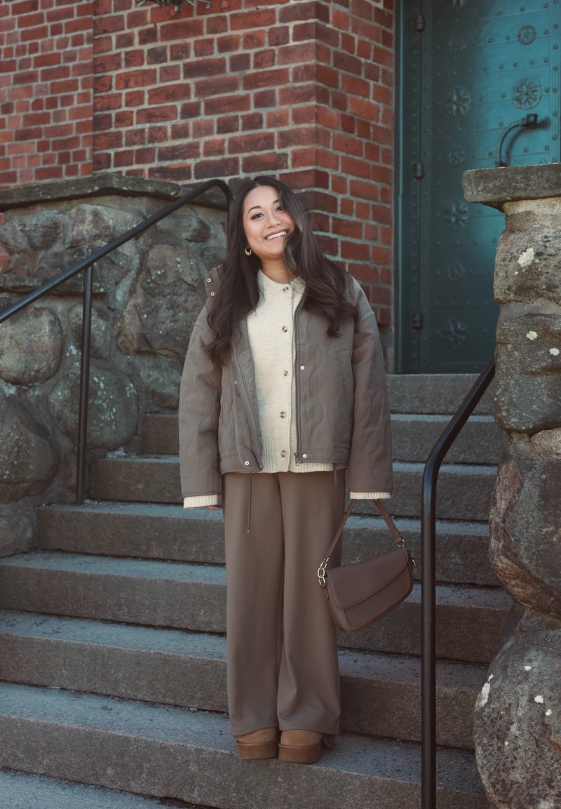 Donna sorridente con abbigliamento casual beige e marrone che tiene una borsa marrone, in piedi su gradini di pietra vicino a un muro di mattoni e una porta verde.