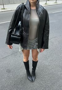 Zwarte leren zip-up jas, doorschijnend grijs topje, grijze rok met studs, en kniehoog zwarte puntige laarzen. Zwarte schoudertas maakt de outfit compleet.
