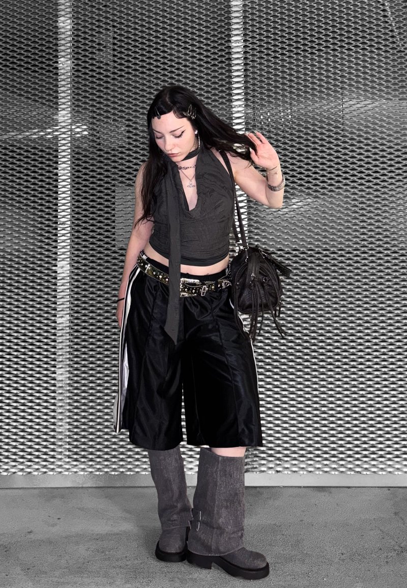 Jeune femme aux longs cheveux foncés portant un haut noir sans manches, un short large, des bottes grises, et tenant un sac noir devant un mur en treillis métallique.
