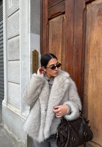 Világosszürke műszőrméből készült kabát, vastag, puha textúrával. Széles gallérral és laza szabással rendelkezik. Barna bőrtáska díszítésekkel.