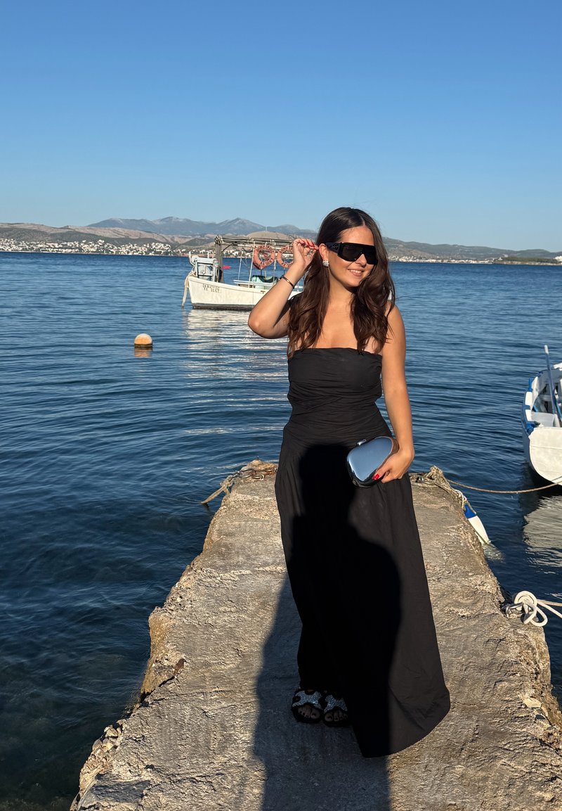 Femme en robe noire et lunettes de soleil debout sur un quai au bord d'une eau calme avec des bateaux et des montagnes en arrière-plan par une journée dégagée.