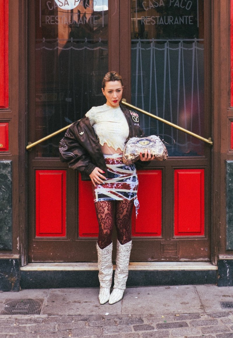 Haut court blanc texturé, veste bomber marron, mini-jupe à motifs rouges et bleus, collants en dentelle noire et bottes étincelantes montant jusqu'aux genoux. Tenant un sac brillant.