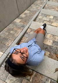 Smiling woman with glasses and headband lying on stone pavement, wearing blue blouse, beige shorts, white socks, and black loafers.