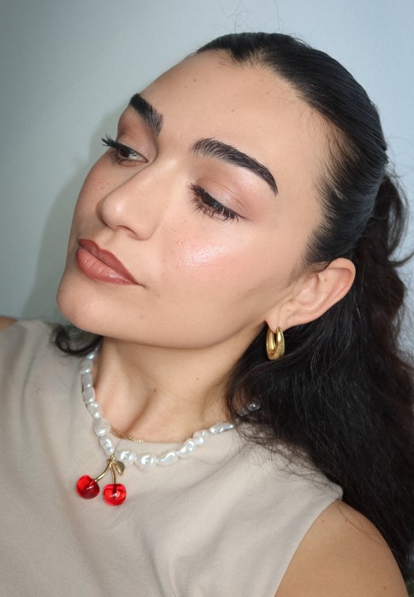 Donna con capelli scuri tirati indietro, indossa orecchini a cerchio dorati, collana di perle con ciondolo a forma di ciliegia rossa, trucco leggero e top beige senza maniche.
