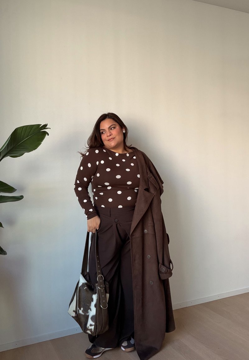 Haut à manches longues marron à pois, pantalon marron à jambes larges, et un long manteau marron ; portant un sac en tissu tacheté. Plante verte en arrière-plan.