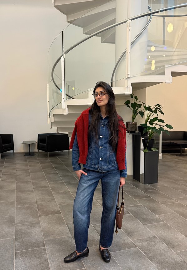 Giubbotto in denim e pantaloni blu scuro, completati da un maglione rosso appoggiato sulle spalle. Mocassini neri e una borsa marrone. Sfondo di interni moderni.