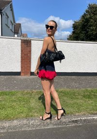 Czarna torebka na ramię o marszczonym wzorze. Modelka w ciemnej bluzce, czerwonej rozkloszowanej spódnicy i czarnych sandałach na obcasie. Jasny, słoneczny dzień.
