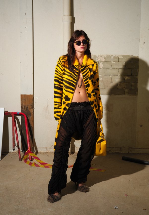 Mujer con abrigo amarillo y negro con rayas de tigre, pantalones negros transparentes, gafas de sol, sosteniendo un bolso amarillo, de pie en un espacio industrial.