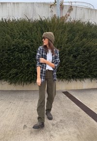 Cămașă în carouri bleumarin și alb peste un tricou alb, pantaloni verde măsliniu, cizme maro și o căciulă maro texturată, pe fundal de verdeață.