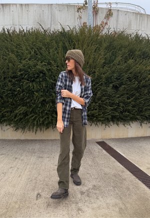 Camisa de cuadros en azul marino y blanco sobre una camiseta blanca, pantalones verde oliva, botas marrones y un gorro marrón texturizado contra un fondo de vegetación.