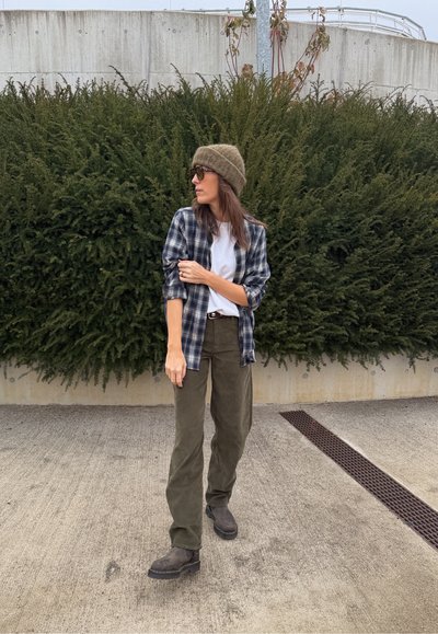 Camisa de cuadros en azul marino y blanco sobre una camiseta blanca, pantalones verde oliva, botas marrones y un gorro marrón texturizado contra un fondo de vegetación.