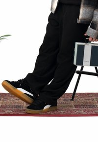 Baskets en daim noir avec des bandes latérales blanches et des semelles en caoutchouc gomme, portées avec un pantalon noir sur un tapis à motif. Radio vintage bleue et argent à proximité.