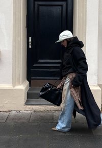 Schwarzer langer Mantel über einem schwarzen Kapuzenpullover, blaue Jeans mit weitem Bein, kariertes Hemd in der Taille gebunden, hält eine schwarze Tasche und trägt eine helle Mütze.