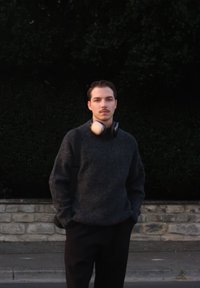 Grå strikket sweater og sorte bukser; iført hovedtelefoner om halsen; står ved en stenmur med grønt i baggrunden.