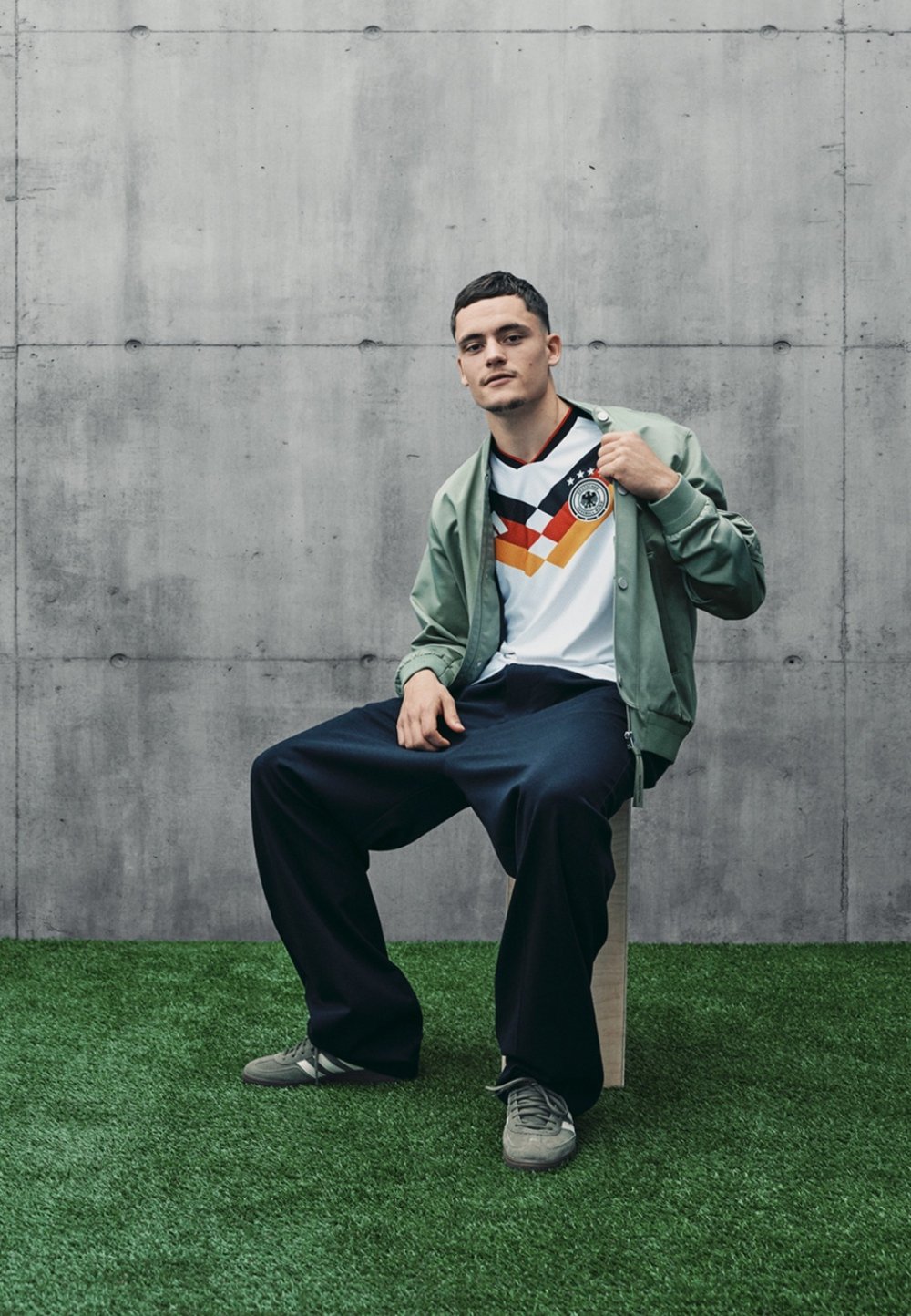 Bílý dres s černo-červeno-žlutou grafikou a logem, v kombinaci se světle zelenou bomber jacket a tmavými volnými kalhotami na umělé trávě.