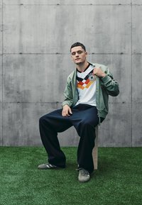 Bílý dres s černo-červeno-žlutou grafikou a logem, v kombinaci se světle zelenou bomber jacket a tmavými volnými kalhotami na umělé trávě.