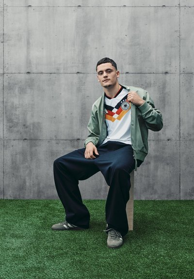Camiseta blanca con gráficos y logo en negro, rojo y amarillo, combinada con una chaqueta bomber verde claro y pantalones oscuros de corte holgado sobre hierba artificial.