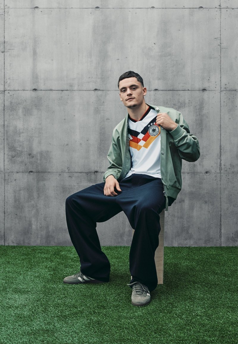 Maillot blanc avec graphiques et logo noir-rouge-jaune, associé à une veste bomber vert clair et un pantalon ample sombre sur de l'herbe artificielle.