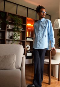 Chemise bleu clair à boutons avec des coutures noires décoratives, accompagnée de pantalons rayés sombres. Le décor comprend des meubles modernes et de la verdure.