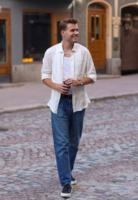 Bílá košile na knoflíky přes bílý top bez rukávů, modré džíny, černé tenisky a tmavý hrnek, prochází se po dlážděné ulici ve městě.