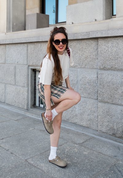 Una camiseta beige clara, una falda de cuadros con tonos azules y verdes, calcetines blancos y zuecos beige con hebillas sobre un pavimento de piedra.