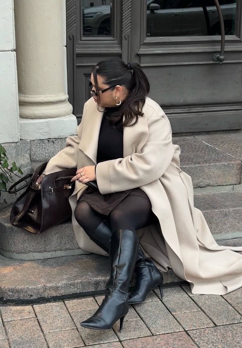 Beige, oversized frakk over en svart topp og brun skjørt. Hun har på seg knelangt, svart støvler med hæler og bærer en mørk brun håndveske.