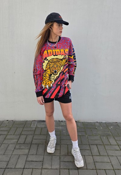 Sudadera de gran tamaño multicolor con un gráfico de tigre y texto "ADIDAS", combinada con pantalones cortos negros y zapatillas grises. Una gorra negra acentúa el estilo.
