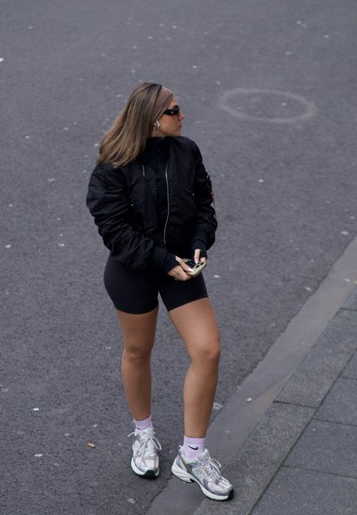 Chaqueta bomber negra, pantalones cortos ajustados negros, zapatillas blancas y grises. Cabello largo, gafas de sol y diadema. Sosteniendo un teléfono. Entorno urbano.
