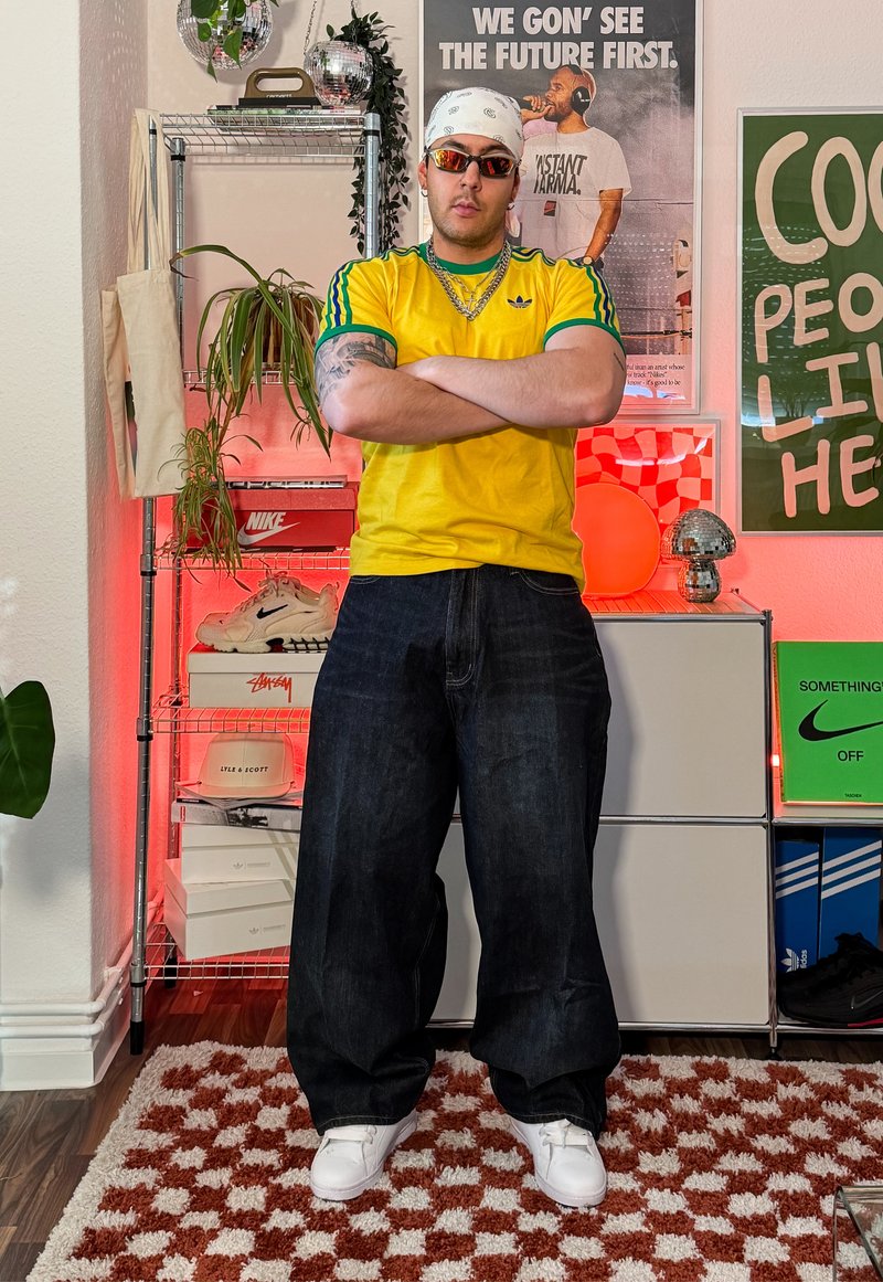 Homme portant des lunettes de soleil et un bandana, bras croisés, vêtu d'un t-shirt jaune Adidas, debout dans une pièce avec des plantes, des affiches, des baskets et un éclairage rouge.