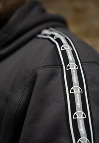 Μαύρη κουκούλα με υφή, που διαθέτει λευκή και μαύρη λωρίδα με λογότυπο κατά μήκος του ώμου, με κυκλικά λογότυπα.