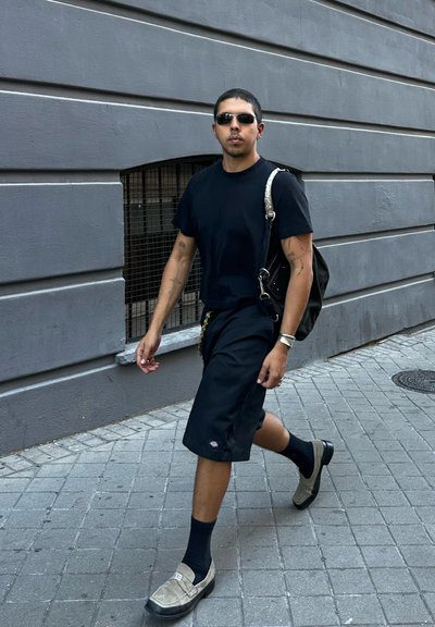 Camiseta negra, pantalones cortos negros hasta la rodilla y zapatos grises sin cordones. Diseño casual con tatuajes visibles en los brazos; llevando una bolsa negra.
