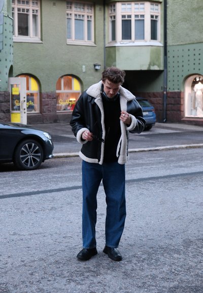 Chaqueta negra forrada de piel de oveja, suéter oscuro, jeans azules y zapatos negros. Entorno urbano con edificios verdes y coches aparcados.