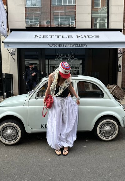 Un coche vintage de color azul claro estacionado en una calle de la ciudad, con una persona que lleva una blusa negra y pantalones blancos fluidos, sosteniendo una bolsa roja.