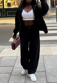 Kvinna som går på stadsgata iklädd svarta byxor, svart jacka, vit Croptop, vita sneakers och bär en liten maroon handväska.