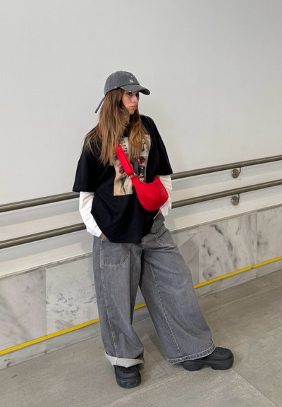 Camiseta negra de gran tamaño con estampado gráfico, superpuesta a una camisa blanca de manga larga. Jeans grises anchos y crocs negros. Bolso cruzado rojo. Gorra gris.