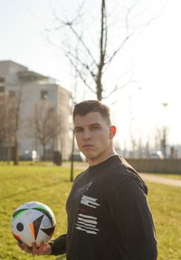 Schwarzes Langarmshirt mit weißen Textdetails; hält einen mehrfarbigen Fußball mit leuchtend grünen, orangefarbenen und blauen Akzenten.