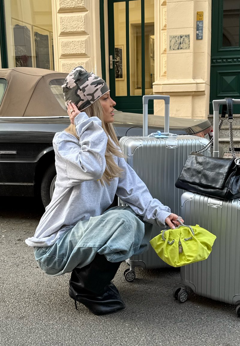 Kvinde sidder på hug ved siden af to sølvfarvede kufferter med hjul, holder en lille gul taske, iført camouflage-beanie, oversized skjorte, løse bukser og sorte støvler.
