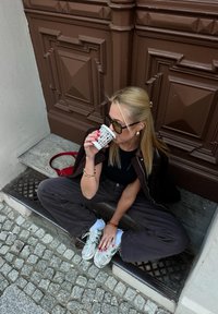 Blond kvinna i solglasögon som sitter på en trappa, klädd i mörka kläder och sneakers, och sippar från en mönstrad papperskopp.