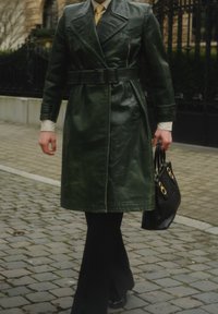 Trench-coat en cuir vert avec une ceinture à la taille, manches longues, et poches latérales, associé à un pantalon noir et un sac noir avec des accents dorés.