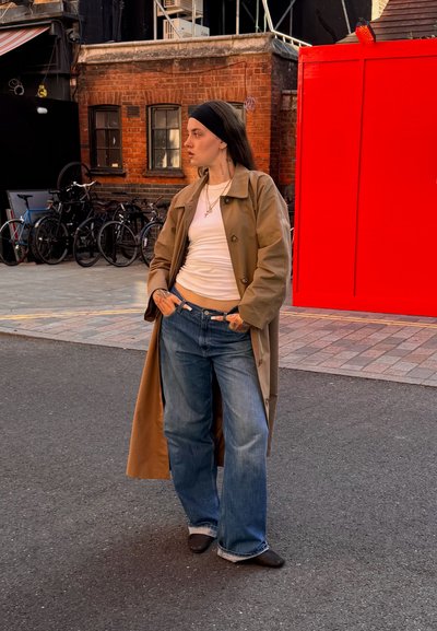 Abrigo trench tan sobre camisa blanca, combinado con jeans anchos azules. Detalles notables incluyen collares en capas y una diadema negra.