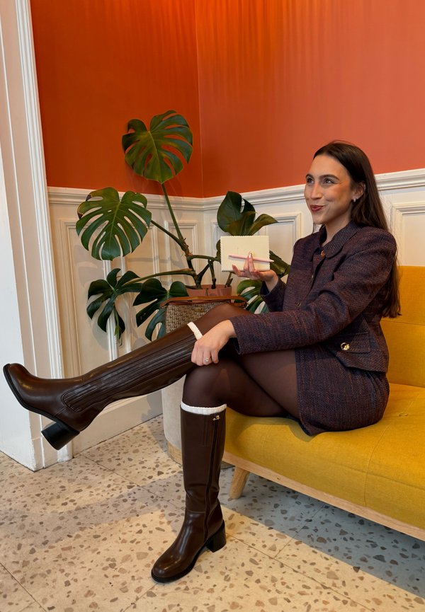 Vrouw met bruine laarzen en bijpassend jack zittend op een gele bank, een klein beige notitieboekje vasthoudend met een roze pen, groene planten in de buurt.