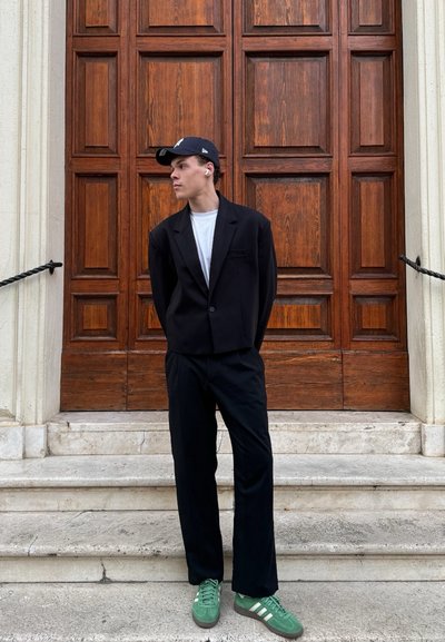 Americana negra sobre una camisa blanca, combinada con pantalones negros. Lleva zapatillas verdes con rayas blancas, de pie contra una puerta de madera.