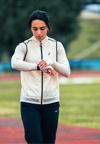 Gilet léger crème avec des accents noirs, porté sur un haut à manches longues. Pantalon foncé, montre-bracelet visible. Fond de piste d'athlétisme en plein air.