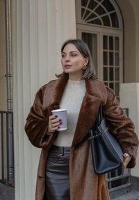 Manteau en fausse fourrure marron, pull côtelé crème, jupe en cuir marron, tenant une tasse de café, avec un sac à main structuré noir et des boucles d'oreilles créoles dorées.