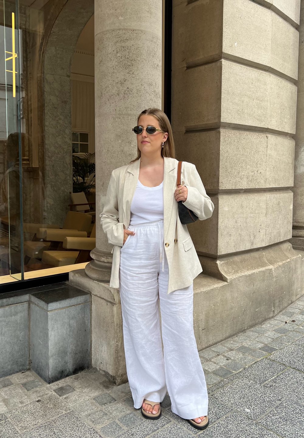 Svijetložuti laneni sako, bijeli top bez rukava i lepršave bijele lanene hlače. Crne sunčane naočale, drži malu torbicu, stojeći pored kamenog stupa.