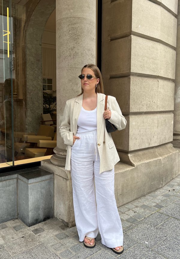 Jasnobeżowa lniana marynarka, biała koszulka na ramiączkach i luźne białe lniane spodnie. Czarne okulary przeciwsłoneczne, trzymając małą torebkę, stojąc przy kamiennej kolumnie.