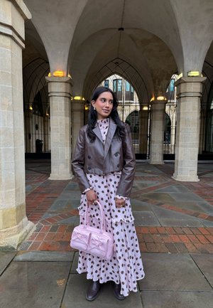 Mujer con chaqueta de cuero marrón y vestido de lunares sosteniendo un bolso rosa, de pie bajo columnas de piedra arqueadas con suelo de baldosas.
